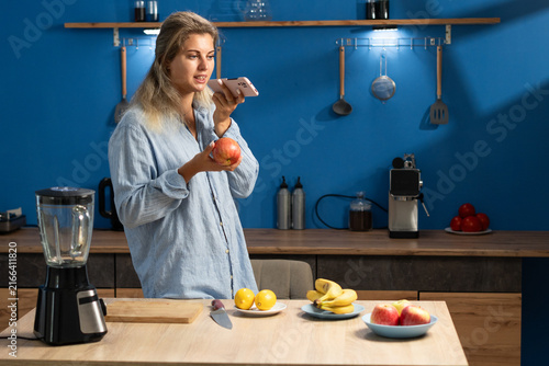 Quadro su tela Woman standing in kitchen holding an apple while dictating a voice message to smartphone