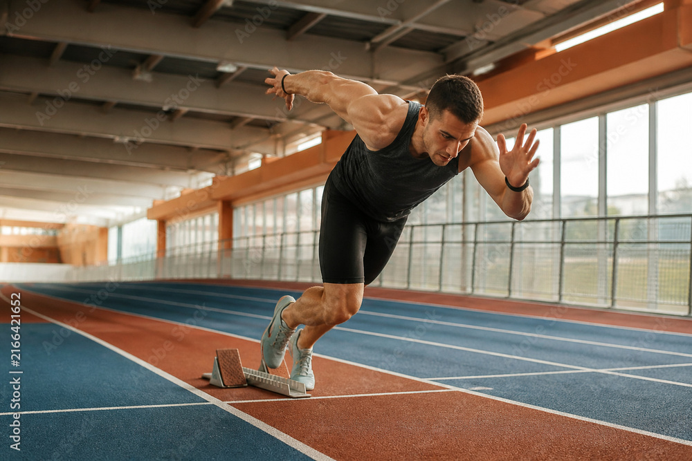 Fototapeta premium Muscular male sprinter starting running race on indoor track. Athletic man in sportswear exploding from starting blocks. Professional sports training concept for speed and power.