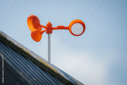Quadro su tela Orange weather station anemometer and wind vane mounted on a rooftop against a c