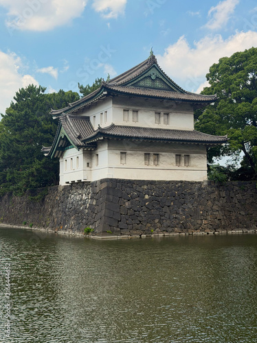 Sakurada-Niju-Yagura Watchtower at the Imperial Palace in Tokyo, Japan, standing above a stone wall beside the moat, surrounded by lush trees under a blue summer sky