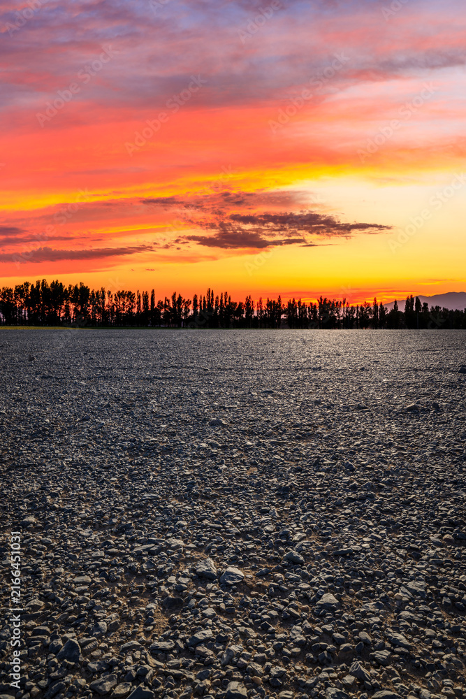 Naklejka premium A vast gravel field and silhouetted trees under a vibrant sunset sky