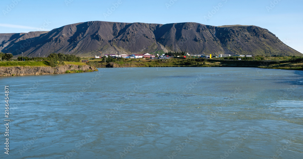 Naklejka premium A scenic view of a wide river flowing through Selfoss with a dramatic volcanic mountain and a small town in the background under a clear blue sky.