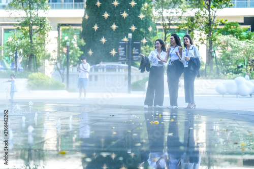 Photography A team of three Hispanic businesswomen in matching dark suits, laughing with heads thrown back, walk across a mirror-like reflecting pool at dusk holding coffee cups on the bench