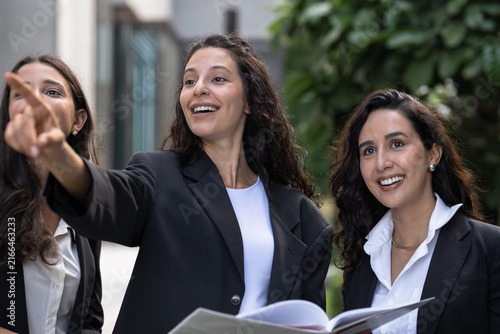 Foto A diverse team of three Hispanic businesswomen in dark suits, wide-eyed at what she sees, share a finding from a printed proposal with a large open report held between them