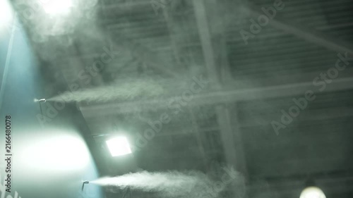 Video shows misty industrial interior with spray jets releasing vapor into air illuminated by bright spotlights. Atmosphere shifts from calm to dense fog as vapor fills space