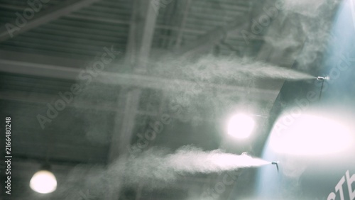 Video shows misty industrial interior with spray jets releasing vapor into air illuminated by bright spotlights. Atmosphere shifts from calm to dense fog as vapor fills space