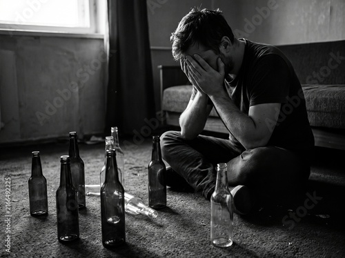 Desperate man holding head in hands surrounded by empty glass bottles