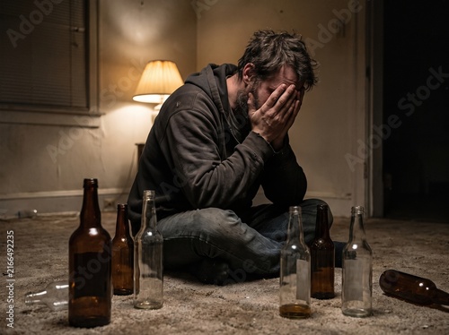 Desperate man holding head in hands surrounded by empty glass bottles