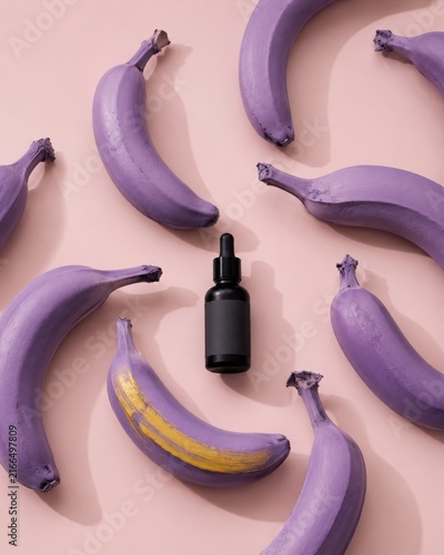 Surreal flat lay composition of purple painted bananas with gold brush stroke surrounding a black dropper cosmetic bottle on pastel pink background creative skincare branding