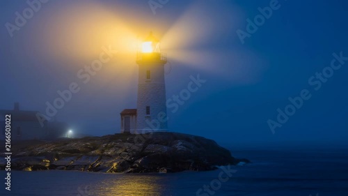 Lighthouse glowing brightly on rocky shore during foggy night ocean scene with calm water