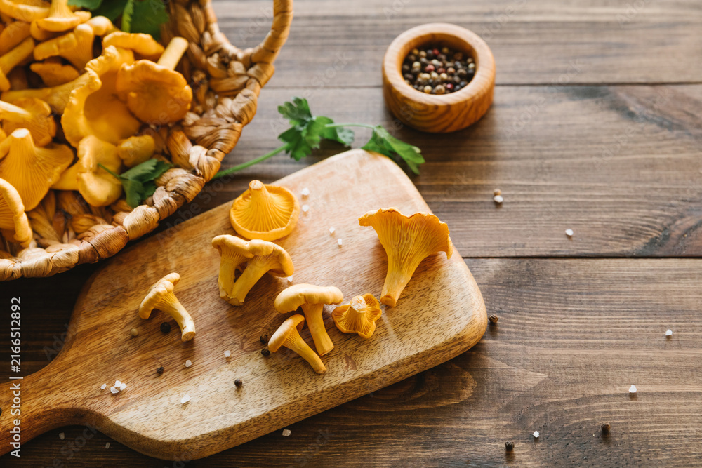 Fototapeta premium Fresh chanterelle mushrooms on a wooden board and in a basket on a dark table, rustic harvest scene, healthy cooking ingredients, organic forest food, autumn season. Top view, flat lay, copy space.