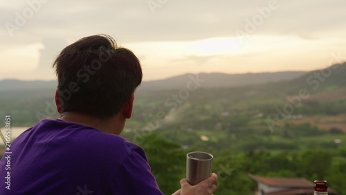 Foto A young man contemplates the future, enjoying a peaceful moment with a drink while overlooking a vast mountain range and valley during a beautiful sunset