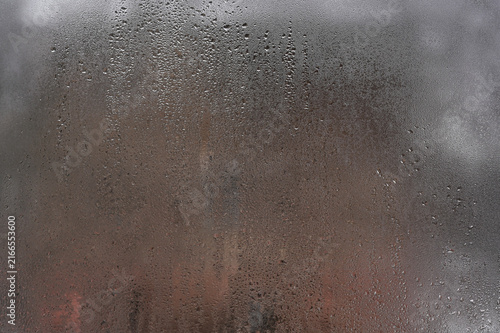 Damp window surface displays thick condensation from indoor temperature contrast. Foggy glass pane accumulates small moisture drops in heated room. Steamy window panel presents soft bokeh background.