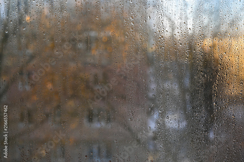 Condensed glass surface shows heavy indoor moisture during chilly weather. Water droplets trickle down misty window pane in cozy home atmosphere. Steamy glass panel captures blurred urban view.