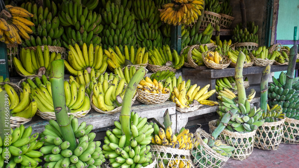 Obraz premium Fresh ripe and green bananas arranged in woven bamboo baskets at a traditional Asian fruit market stand. Organic tropical harvest stacked neatly on wooden shelves for local street market sale.