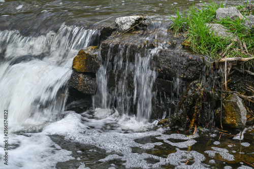 Stream pours across stone cascade beside natural riverbank with bubbling water texture. Waterway highlights peaceful nature retreat and scenic travel inspiration for hikers and explorers.