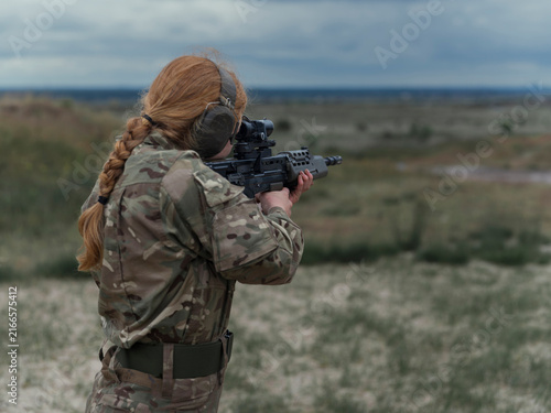 Fotografie Close view of a red haired servicewoman in camouflage aiming an SA80 rifle downrange, ear defenders and safety glasses correctly worn