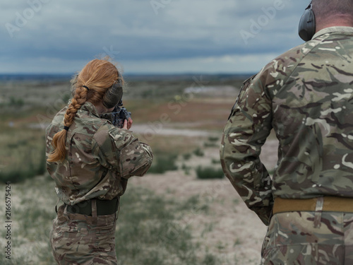 Принти на полотні A female soldier fires from the standing position while a senior soldier watches her technique, hands clasped behind his back