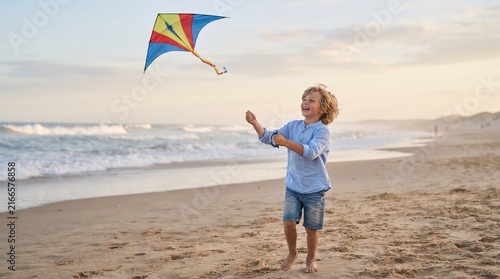 Little boy flying a colorful kite barefoot on a sandy beach during sunset.