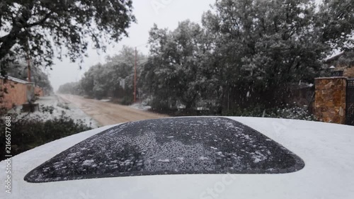 Slow motion snowfall over a snow covered car roof beside an unpaved road lined with trees