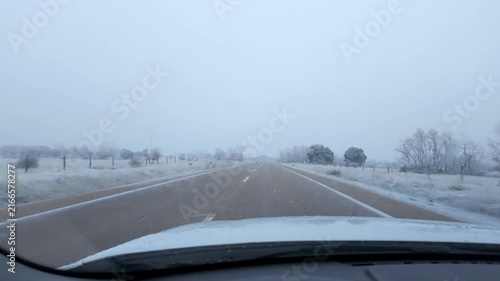 Vehicle point of view along a snowy rural road with frosted trees and falling snow in winter