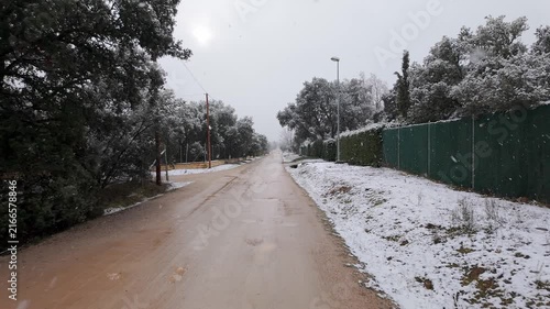 Snowy rural dirt road bordered by trees fences and street lights during steady winter snowfall