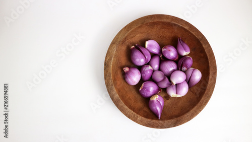 Fresh red shallots are arranged on a rustic wooden plate against a clean white background with copy space