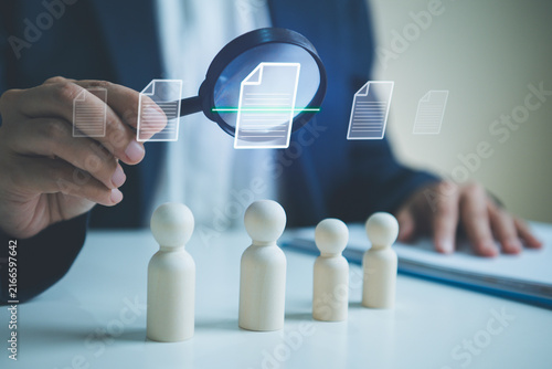 Fotografie Candidate screening with magnifying glass and wooden figures businessperson reviewing digital documents to identify qualified talent improve recruitment decisions and build a skilled workforce