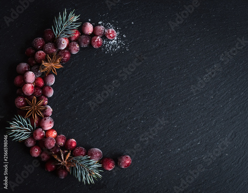 Frosted cranberries, star anise, and pine sprigs form a beautiful crescent shape on a textured dark background, evoking a festive winter mood