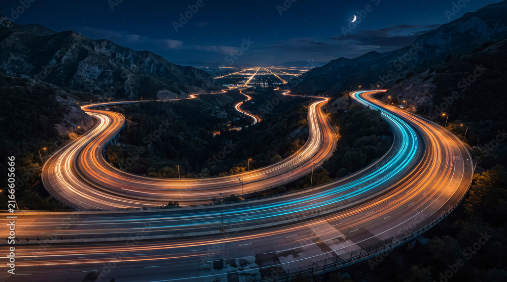 Fototapeta premium Panoramic highway winding through mountain landscape with vibrant light trails at night for infrastructure and travel marketing.