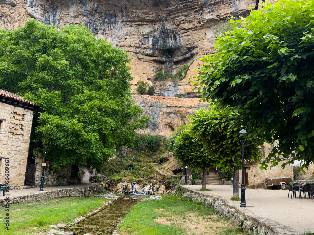 Fototapeta premium peaceful still scene of a small waterfall in a natural landscape with rocky cliff face and trees, captured from an eye-level perspective in an outdoor park or garden area with stone pathway.