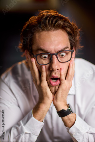 Young man in white shirt with glasses expressing shocked surprise holding face in hands under warm studio light, with man, young, surprised, shocked detail, showing expression, glass, hand, face