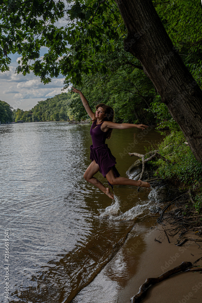 Fototapeta premium Young Woman Jumping Into River From Tree-Lined Shore While Splashing Water In Summer