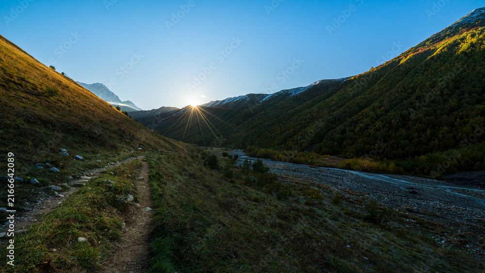 Obraz premium Landscape photograph of sunrise with sunstar flare over mountain valley and hiking path near Adishi Glacier in Svaneti Georgia.