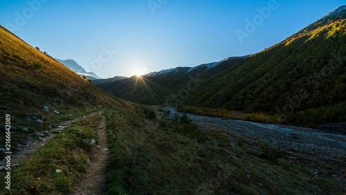 Landscape photograph of sunrise with sunstar flare over mountain valley and hiking path near Adishi Glacier in Svaneti Georgia.