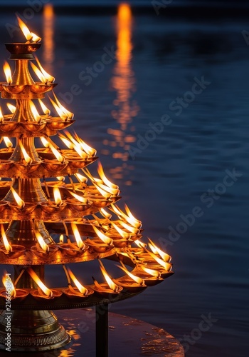 Golden tiered oil lamps flicker with bright orange flames during a spiritual night ritual, casting a warm glow over peaceful river water, light, ancient, water