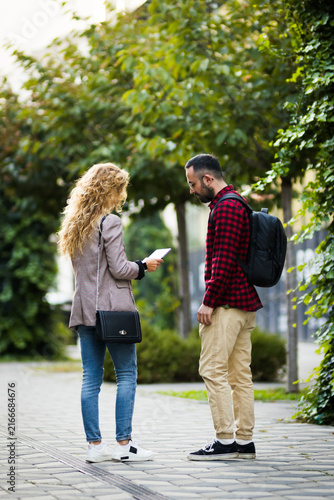 Картината върху платно Young couple standing on a city sidewalk, engaged in conversation while facing each other, surrounded by greenery and trees