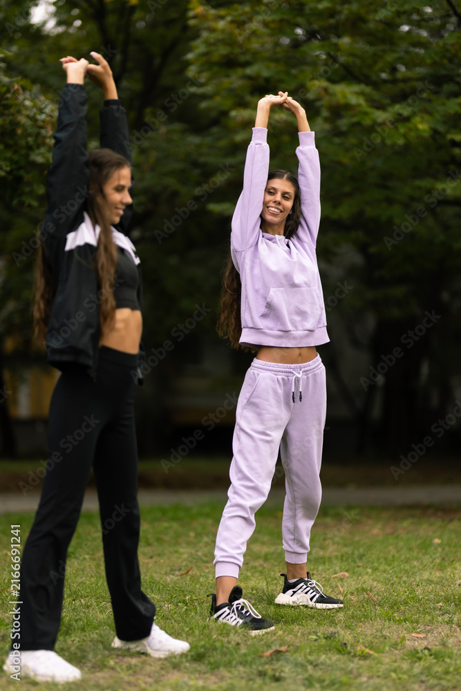 Fototapeta premium Two women stretch together in a park during a sunny day while wearing athletic clothing and enjoying their fitness routine
