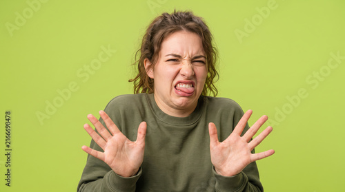 Disgusted woman sticking out tongue with hands up on green background.