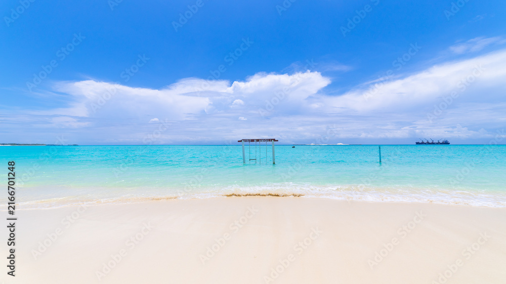 Fototapeta premium Wooden swing standing in the shallow turquoise lagoon on Maamigili Island with gentle waves and dramatic clouds over the Indian Ocean.