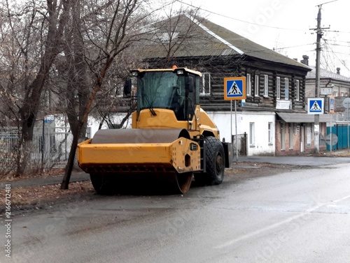 A yellow road roller parked on a wet street next to old houses.