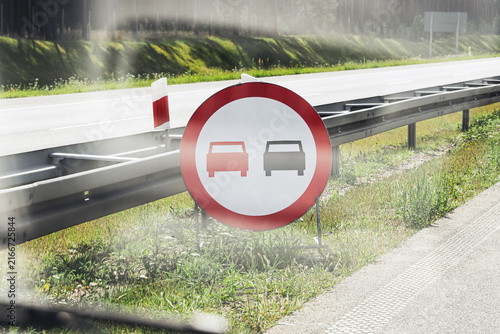 Road sign prohibits vehicle overtaking. Red border marks traffic restriction. Highway barrier runs beside roadway. Driving rules support safer travel. European road infrastructure forms background.