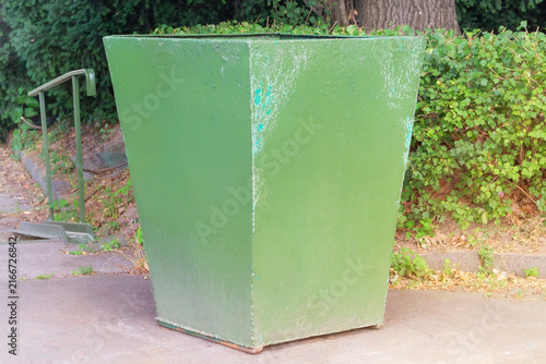 Green metal trash can in city park. Garbage, waste lots, junk dump. Dumpsters being full with garbage. Sunny day.