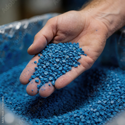 A close-up view of a hand holding a handful of small blue plastic regrind pellets over a container full of processed recycled plastic material.