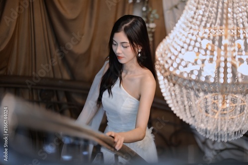 Elegant young woman in white one-shoulder gown descending staircase beside crystal chandelier in luxury interior