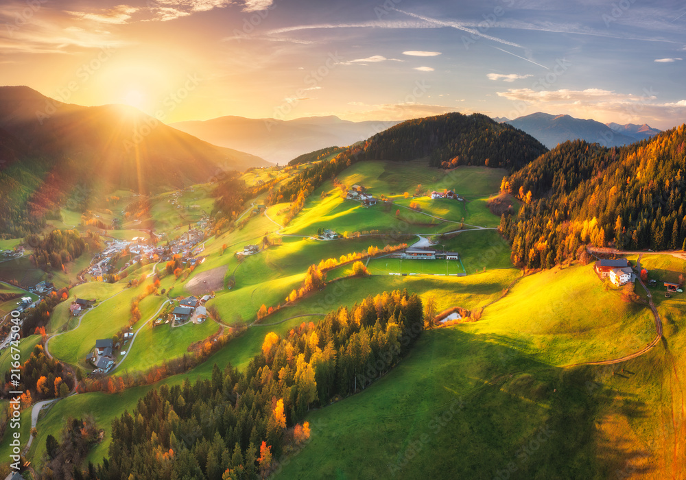 Fototapeta premium Santa Magdalena village valley with green fields in Dolomites, Italy at sunset. Aerial drone view of alpine autumn landscape with sunbeams, forest, houses, rolling hills and mountain range. Top view