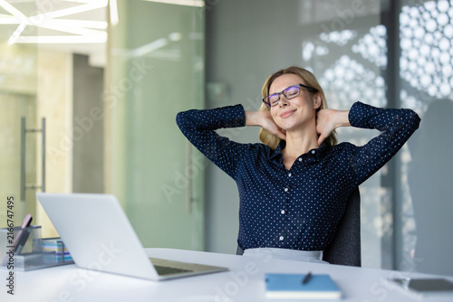 Businesswoman relaxing at her modern office desk  stretching arms behind head and smiling with closed eyes  finding relief from stress during a well-deserved work break