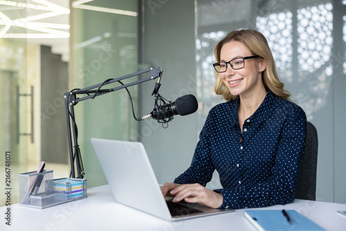 Obraz na płótnie Woman in glasses smiling and typing on a laptop, using a professional microphone