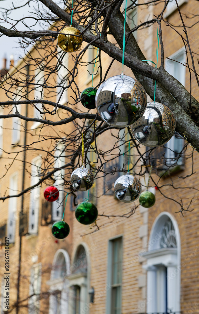Fototapeta premium Decorative baubles hanging from tree branches in front of terrace of Georgian houses in Dublin city. Outdoor exterior ornaments or decorations for Christmas Holiday season. Ireland