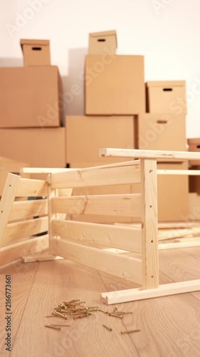 Light wood chair frame lies on wooden floor. Scattered screws surround the frame. Cardboard boxes stack in the background. Furniture assembly work is underway